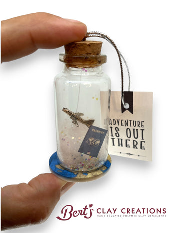 Decorative bottle with cork stopper, airplane, passport, and 'Adventure is Out There' tag, held by a hand with 'Bert's Clay Creations' branding.